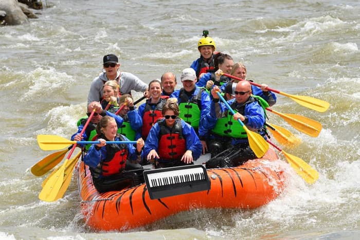 Group in an orange raft with a keyboard strapped to the front paddling through rapids on the Yellowstone River