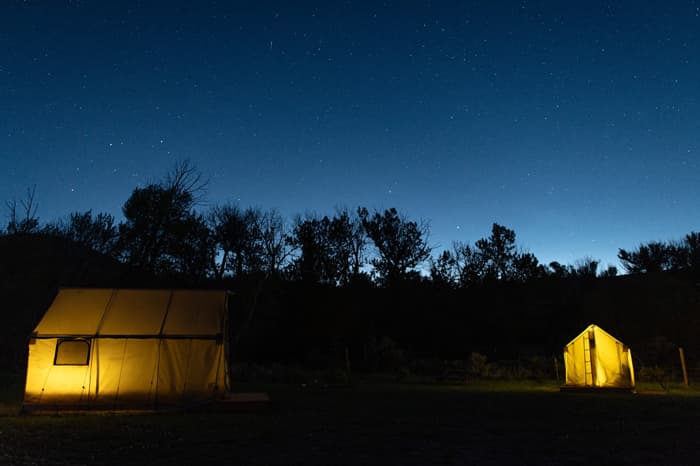 Two canvas tents lit up against a dark forest under the night sky